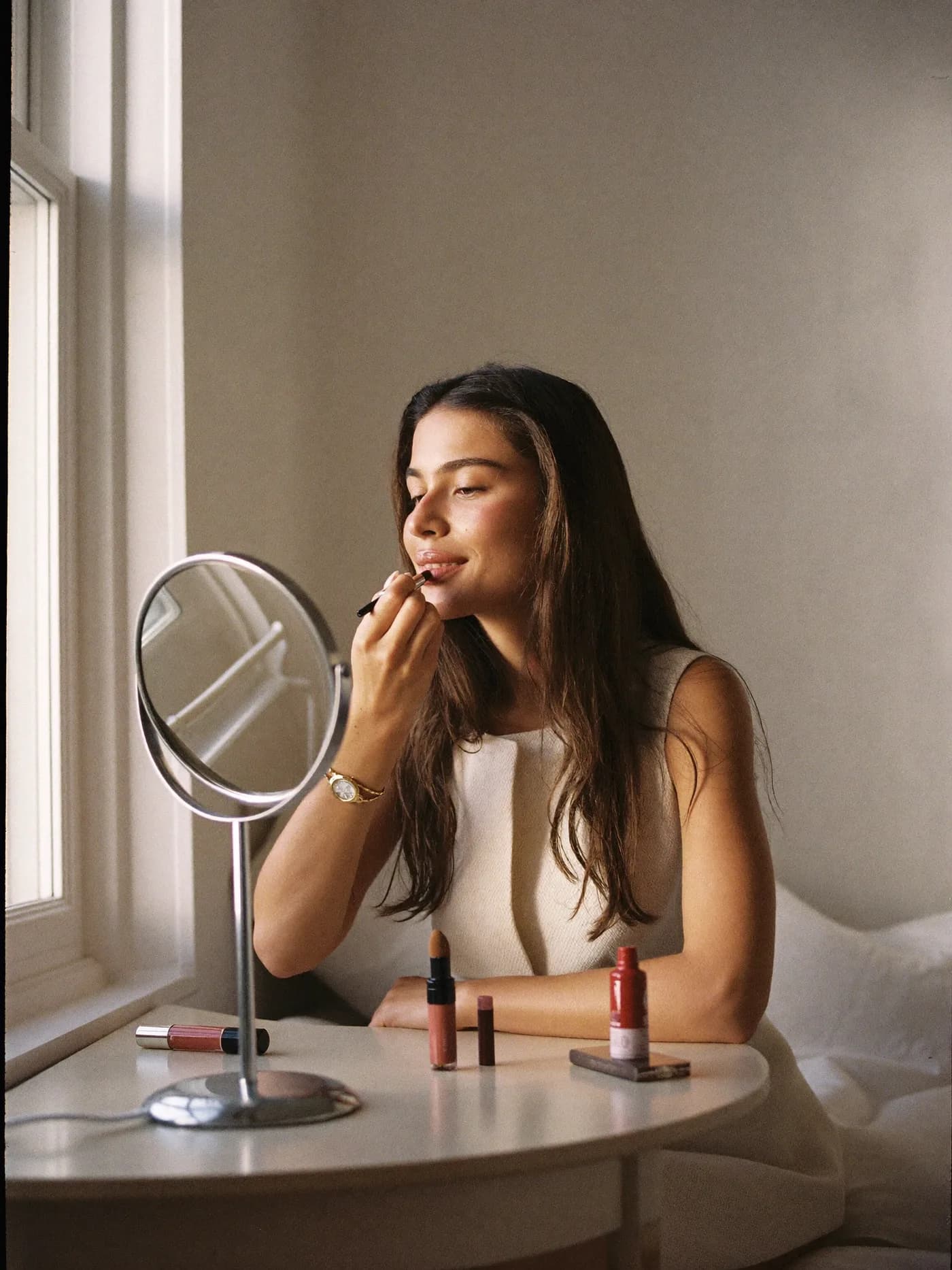 Una mujer se pinta los labios frente a un espejo de mesa en su cuarto, con la luz de la ventana entrando de lado y sus productos de maquillaje sobre la mesa.